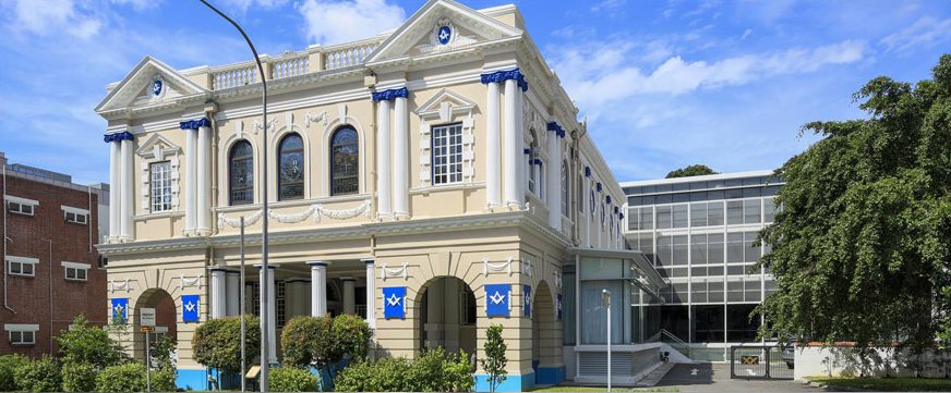 Freemasons’ Hall, Singapore: a two-storey classical building with pedimented bays, engaged columns and arched windows, masonic banners hung between the columns, and a glazed four-storey annexe behind it.