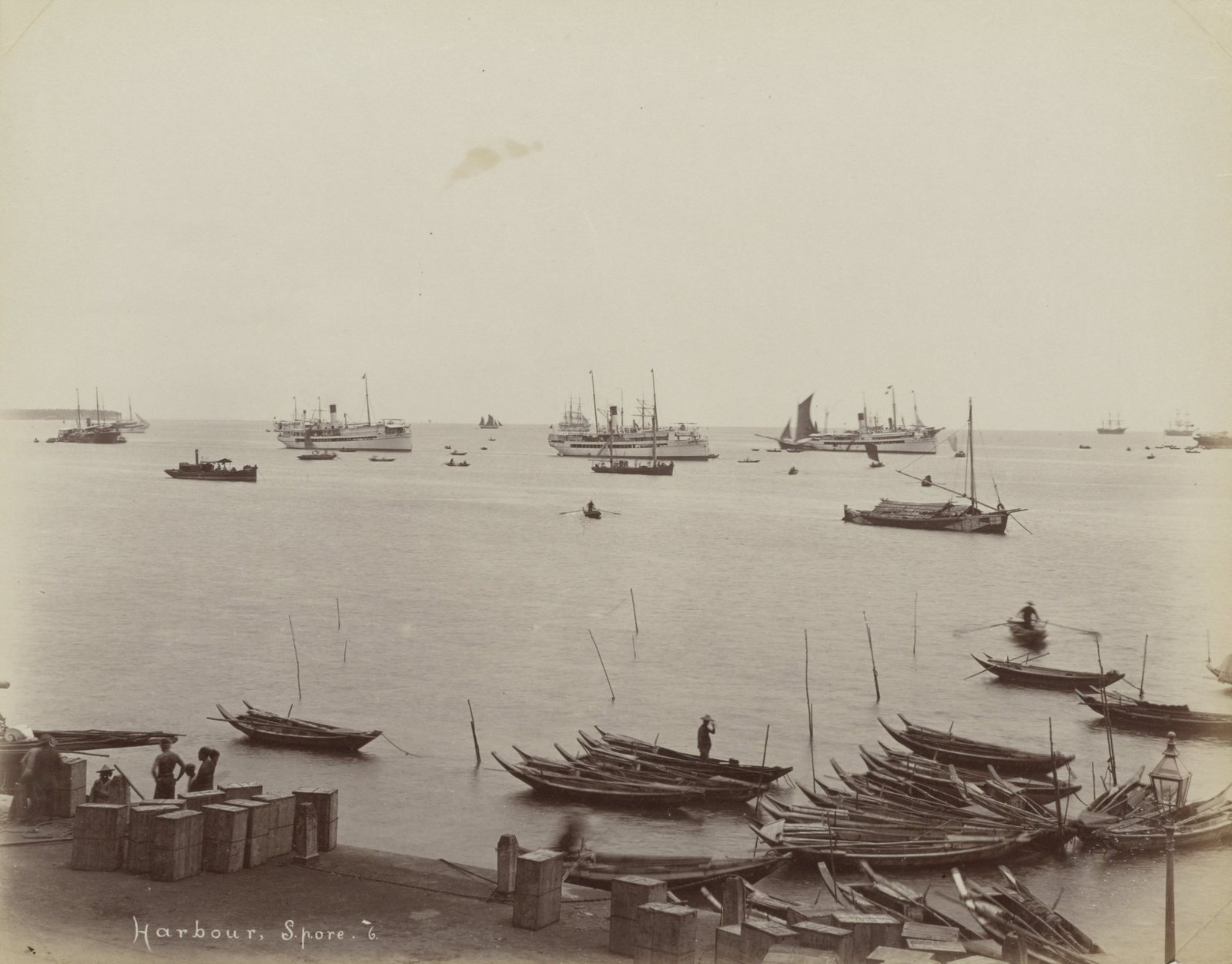 Singapore harbour in the late nineteenth century: steamers and sailing ships lying at anchor in the roads, with sampans drawn up along the shore and cargo cases stacked on the quay in the foreground.